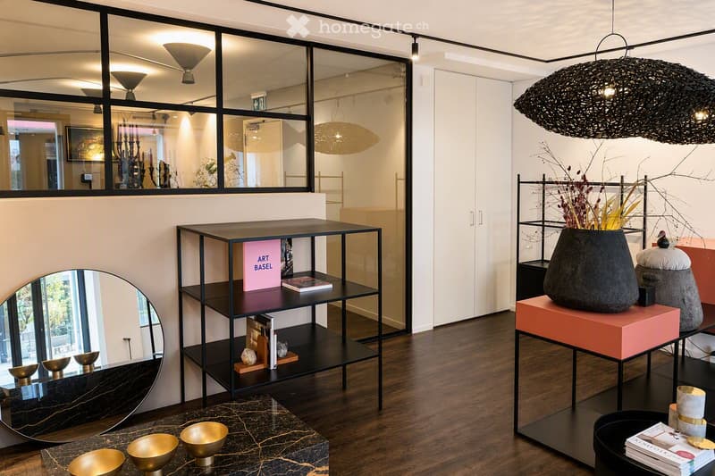 Modern interior with dark wooden flooring, large mirror, black shelving unit holding books and decorative items, a large vase on a pink stool, and several golden bowls on a dark table.
