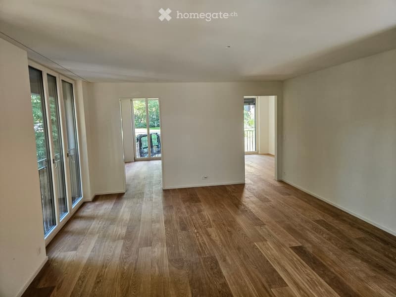 Empty room with light wooden flooring, white walls, large glass sliding doors leading to a balcony, and a view of greenery outside.