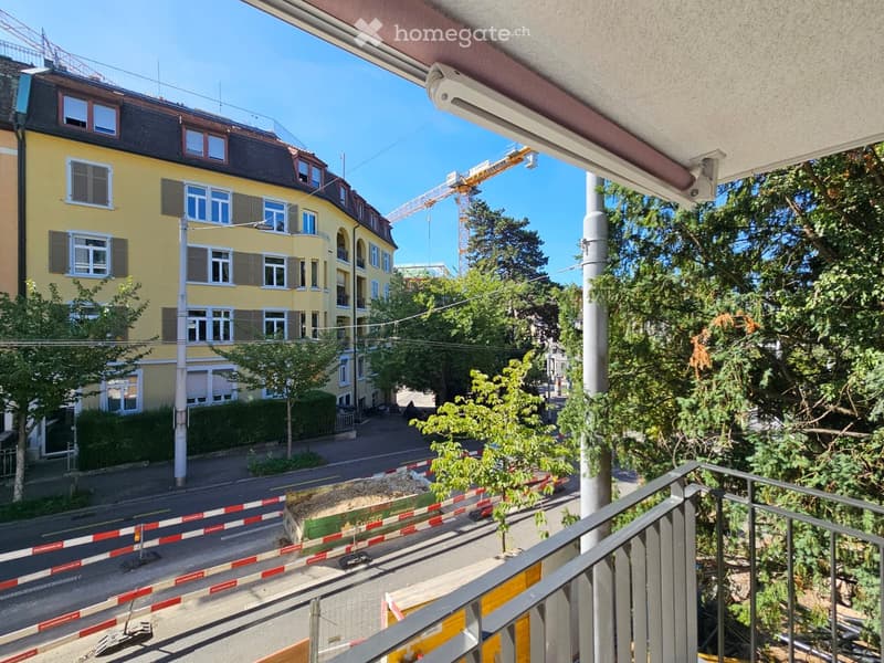 Balcony overlooking a street with a yellow building, trees, and construction barriers.