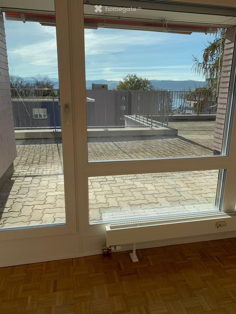 View from inside a room showing a balcony with a tiled floor, metal railing, and scenic outdoor view of water and mountains.