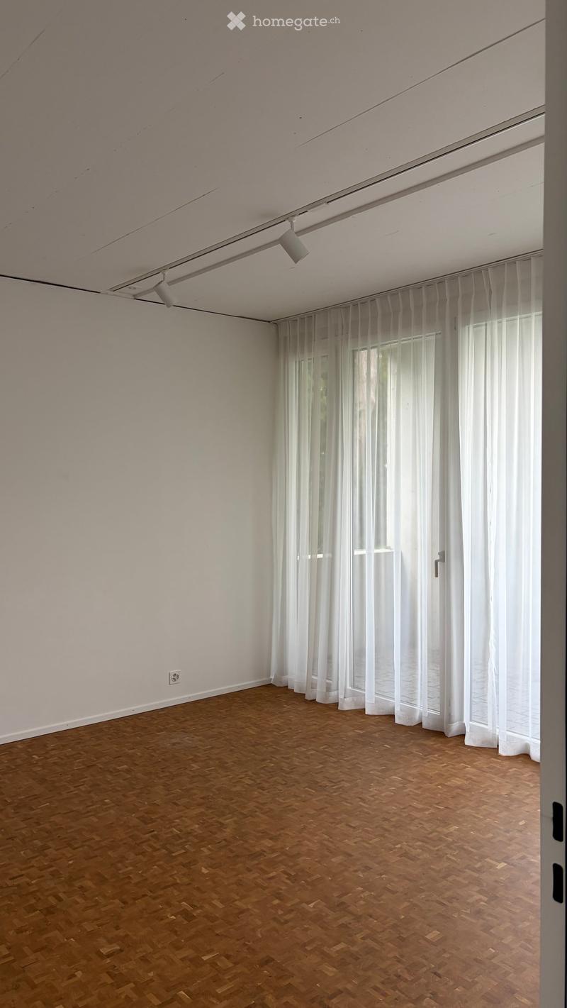 Empty room with wooden parquet flooring, white walls, large windows with sheer white curtains, and recessed ceiling lighting.