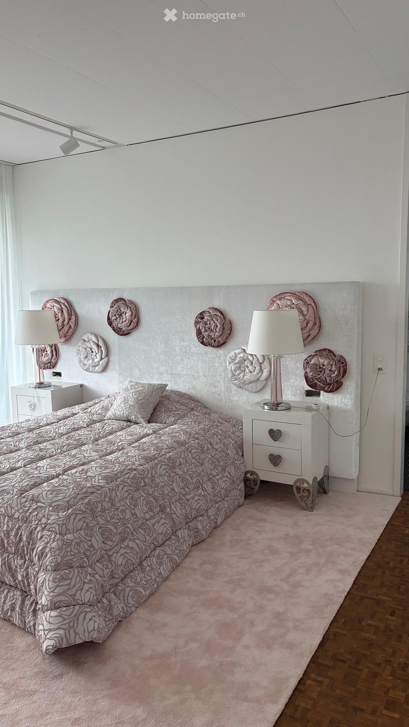 Elegant bedroom with a large bed, matching nightstands, lamps, and decorative plates on the headboard wall. The room features a pink carpet and wooden flooring.