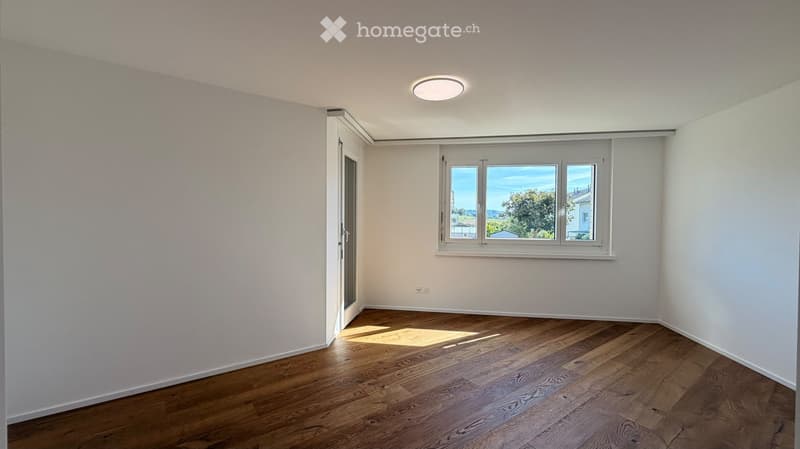 Empty room with wooden flooring, white walls, and large windows offering a view of the outdoors.