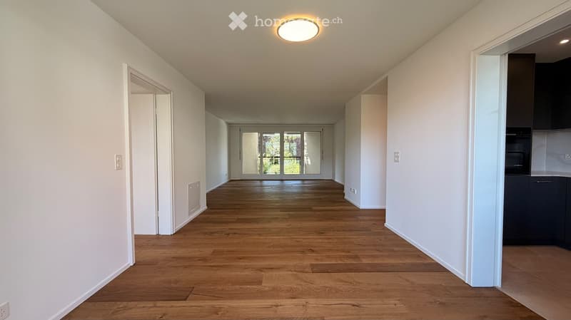 Empty hallway with wooden flooring, white walls, and recessed lighting. Features an open doorway leading to a room with large glass doors and a glimpse of a kitchen area.
