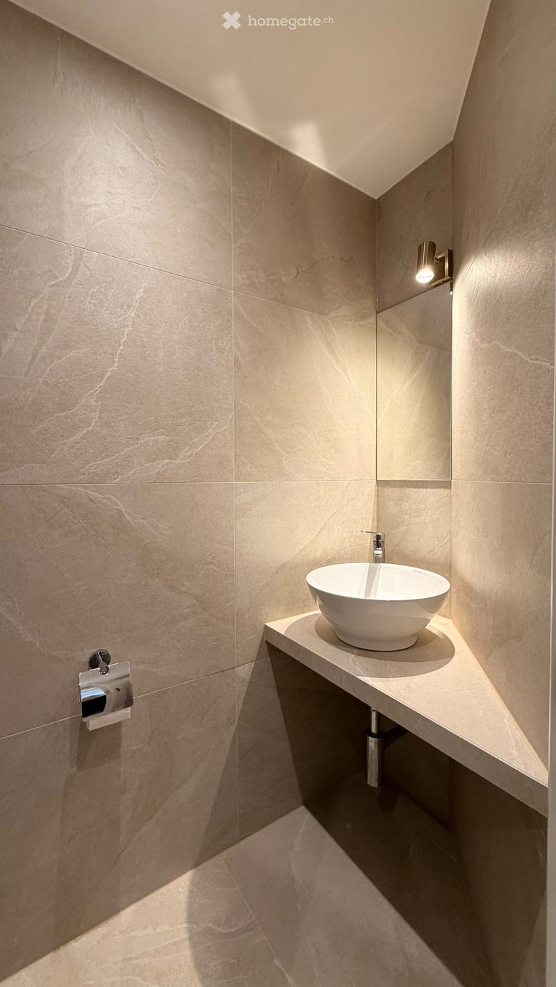 Modern bathroom with a sleek, minimalist design featuring a white sink, chrome faucet, and a wall-mounted toilet paper holder. The walls and floor are covered in light beige marble tiles, creating a clean and contemporary aesthetic.