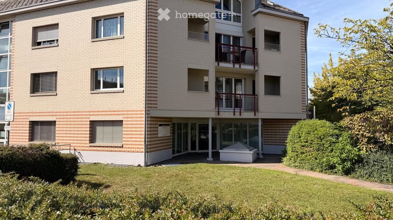 A modern multi-story apartment building with beige brick facade, large windows, and balconies. The ground floor features a glass entrance and a small garden area with neatly trimmed bushes and a small white structure.