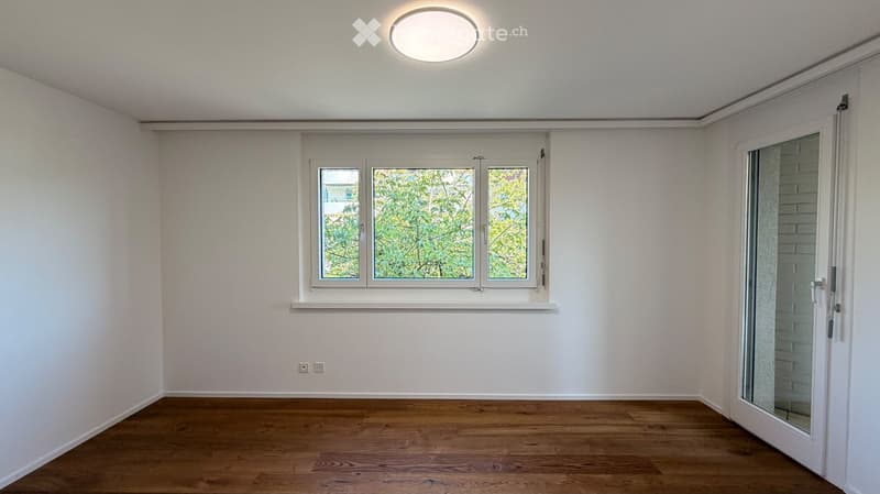 Empty room with light-colored walls, wooden flooring, a large window with three panes, and a sliding glass door leading outside.