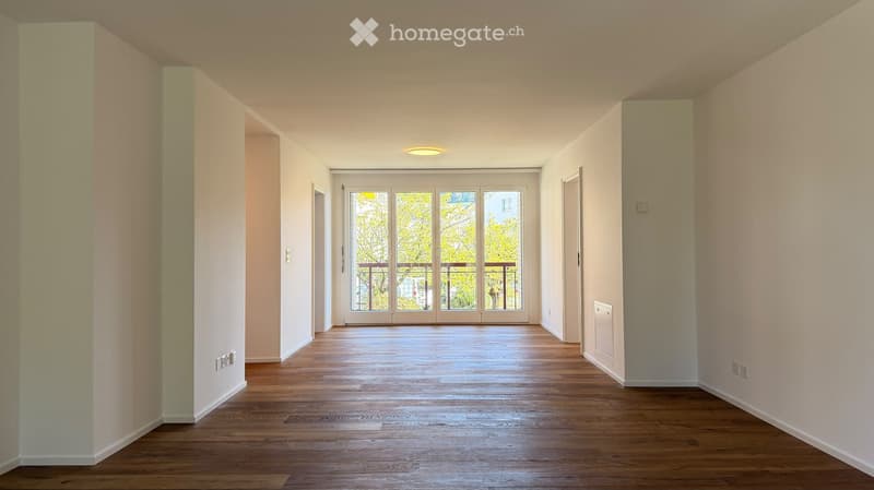 Empty hallway with wooden flooring, white walls, and a large glass door leading to a balcony area.