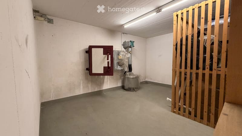 Empty utility room with concrete floor, white walls, fluorescent lighting, and a wooden barrier partially enclosing the space. Includes a fire alarm box and a covered appliance on the floor.