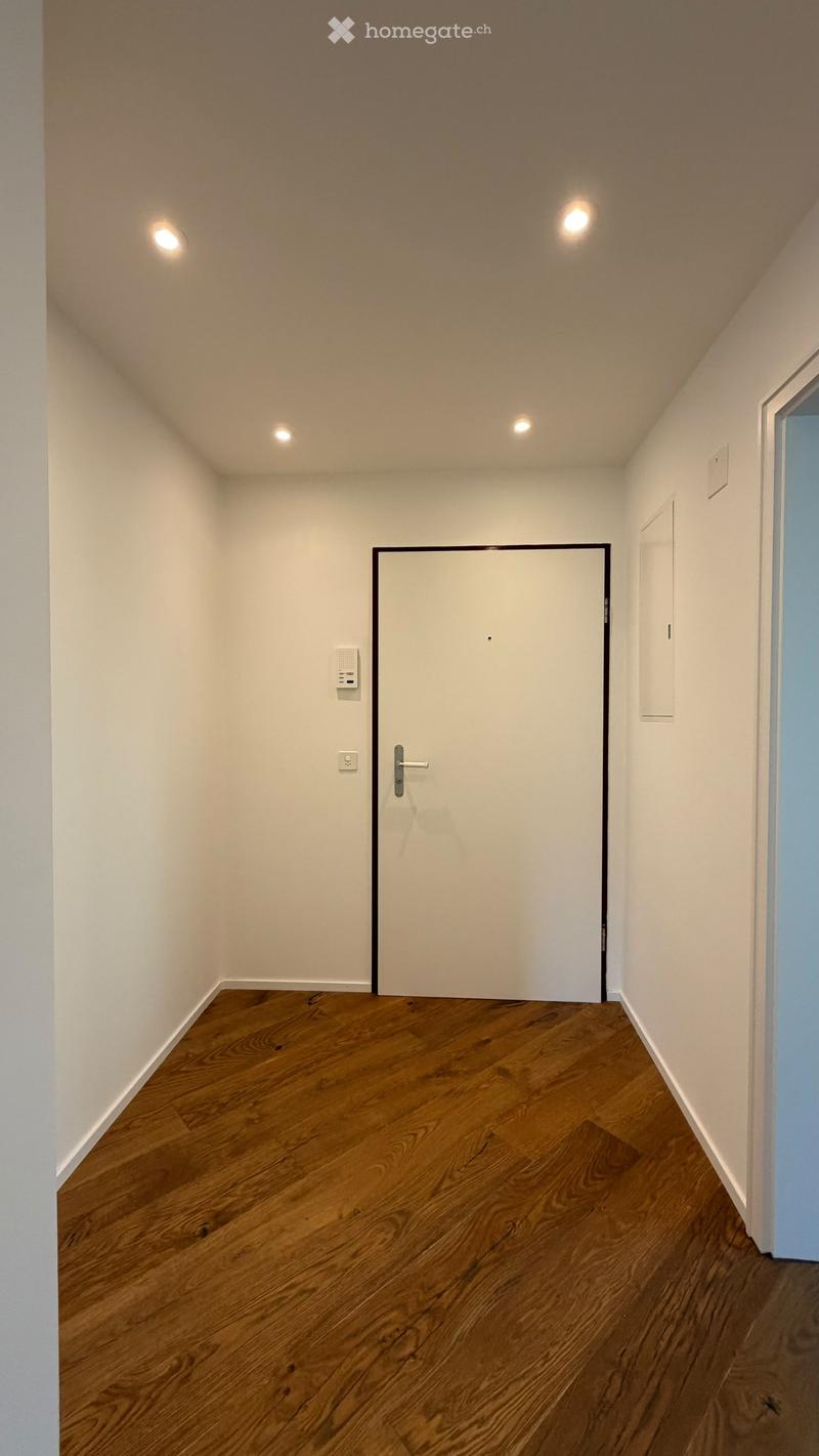 Empty hallway with wooden flooring, white walls, and recessed lighting. A closed door is visible in the center.