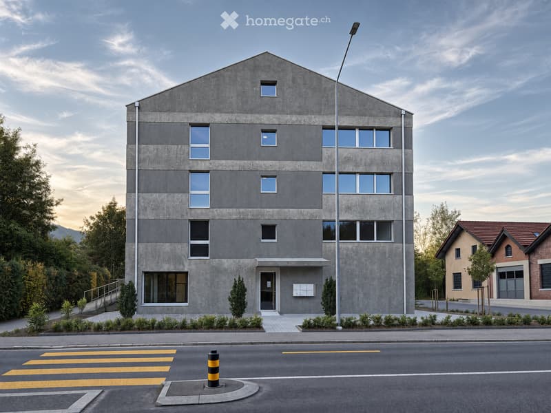 Modern two-story building with grey concrete facade, multiple windows, and a small entrance. The building is surrounded by greenery and a street with a pedestrian crossing.