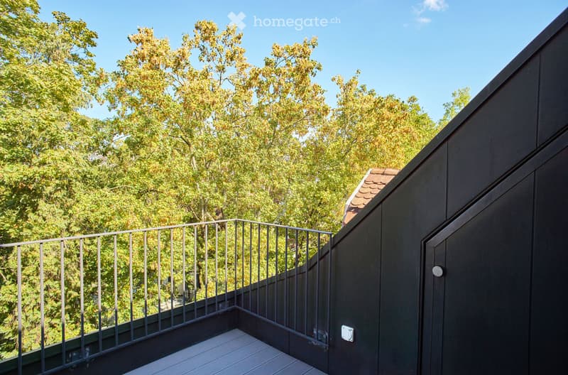 A modern balcony with a metal railing, wooden decking, and lush green trees in the background.