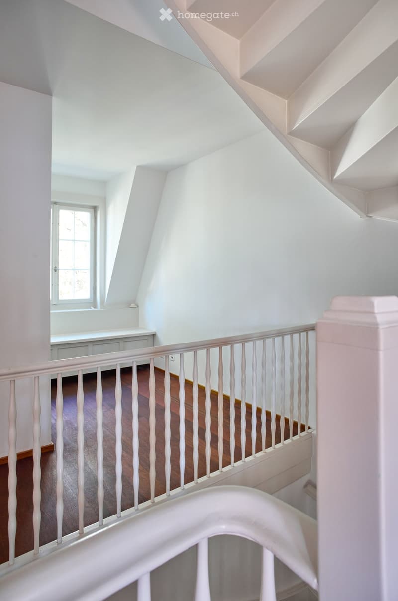 A modern, well-lit staircase with white walls, wooden steps, and a white balustrade. The landing features a window and a small built-in bench or shelf.
