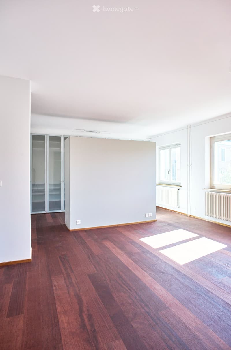 Empty room with wooden flooring, white walls, large windows, and a sliding closet door.