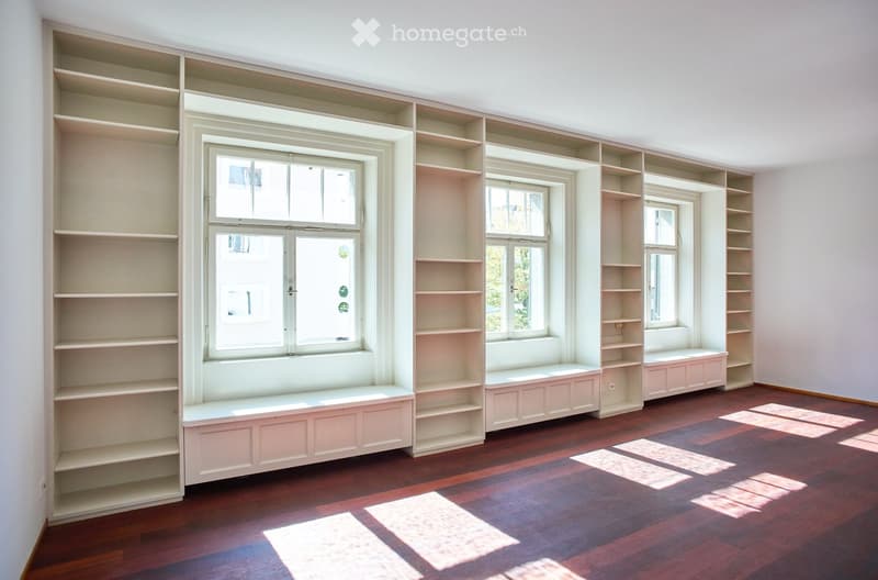 Empty room with built-in shelves and window seats, wooden flooring, and natural light from multiple windows.