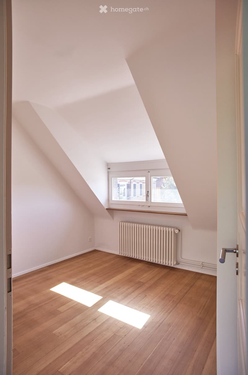 Bright attic room with wooden flooring, white walls, and a radiator. Features a skylight and a window with a view of the outside.