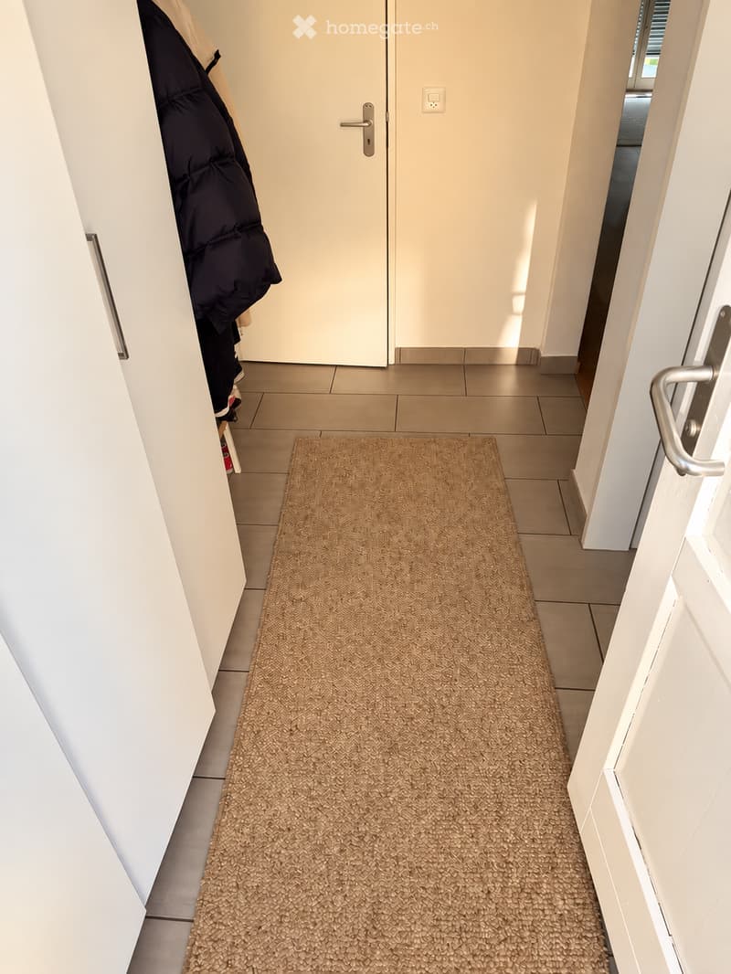 A hallway with tiled flooring, a natural fiber doormat, white walls, and a closed white door with a silver handle. A black puffer jacket is hanging on the left side.