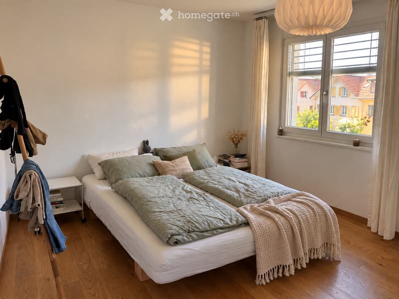 A well-lit bedroom with a large bed, green bedding, a nightstand with books and a vase, and a coat rack with jackets. The room features wooden flooring and large windows with curtains.