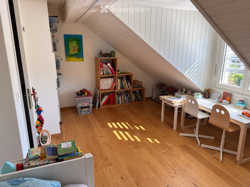 Bright attic room with wooden flooring, slanted ceiling, and large windows. Contains a bed, desk with chairs, bookshelf, storage boxes, and various toys and books.