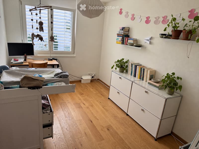 A well-lit room with wooden flooring, a desk with a computer and chair, a cabinet with books and plants, and a toy mobile hanging from the ceiling.