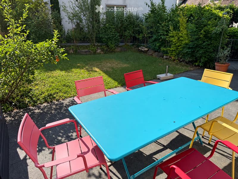 Garden patio with a turquoise table and colorful chairs, surrounded by greenery and a white building in the background.