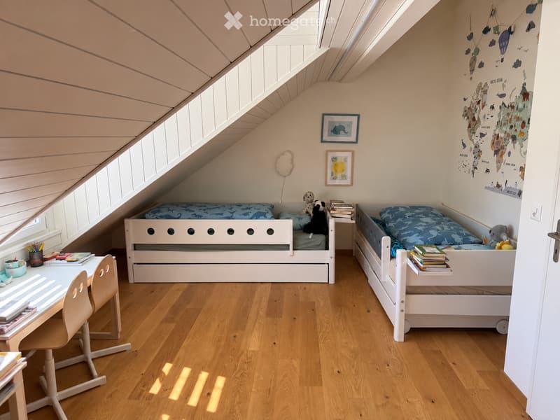 Cozy attic bedroom with slanted wooden ceiling, two single beds with blue bedding, a small desk with chair, and decorative wall art.