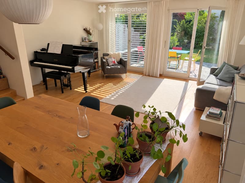 Bright living room with wooden flooring, large windows, and a view of the garden. Features a sofa, armchair, dining table with chairs, piano, and decorative plants.