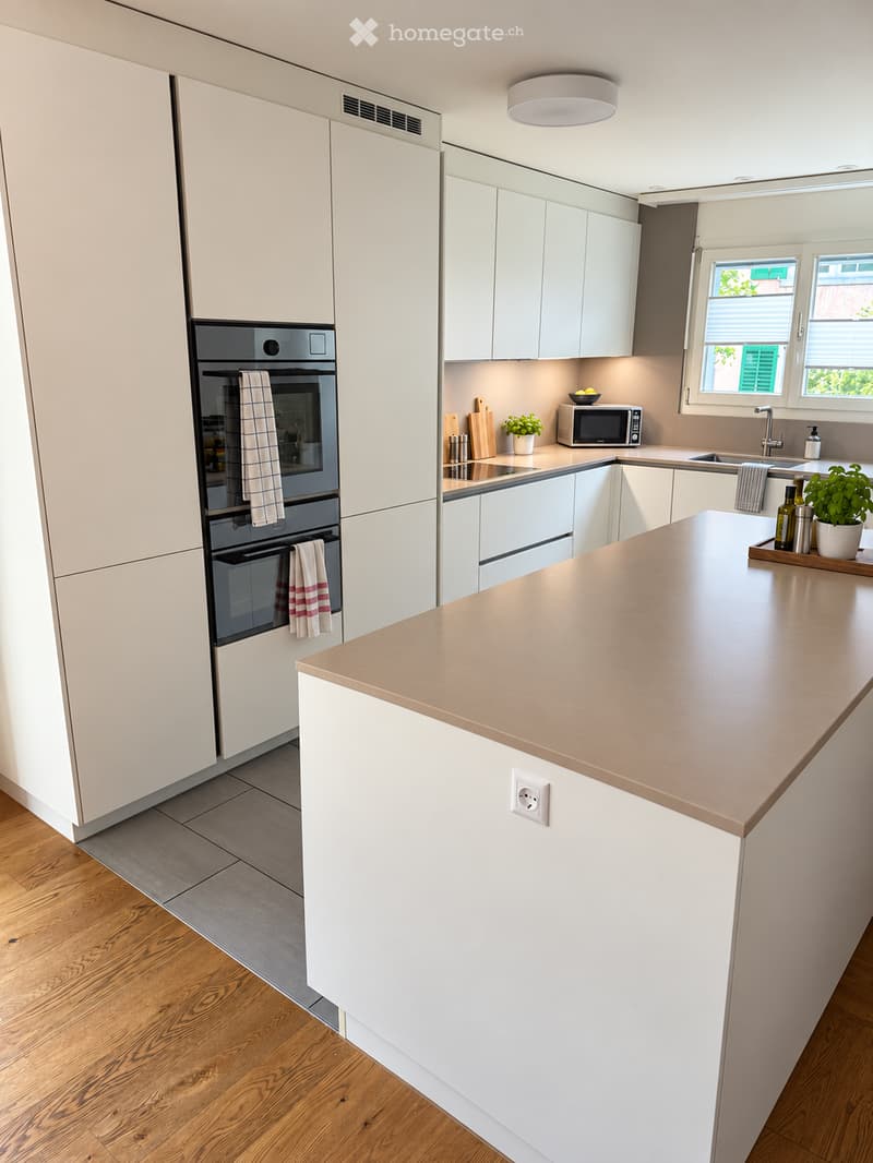 Modern kitchen with white cabinets, light wood flooring, and a central island with a beige countertop. Features include an oven, microwave, sink, and small potted plants on the counter.