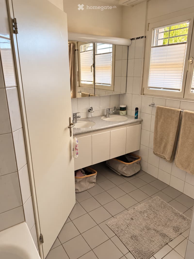Modern bathroom with white tiled walls and floor, double sink vanity with mirrored cabinet, two windows with blinds, towels hanging on a rack, and a small rug on the floor.