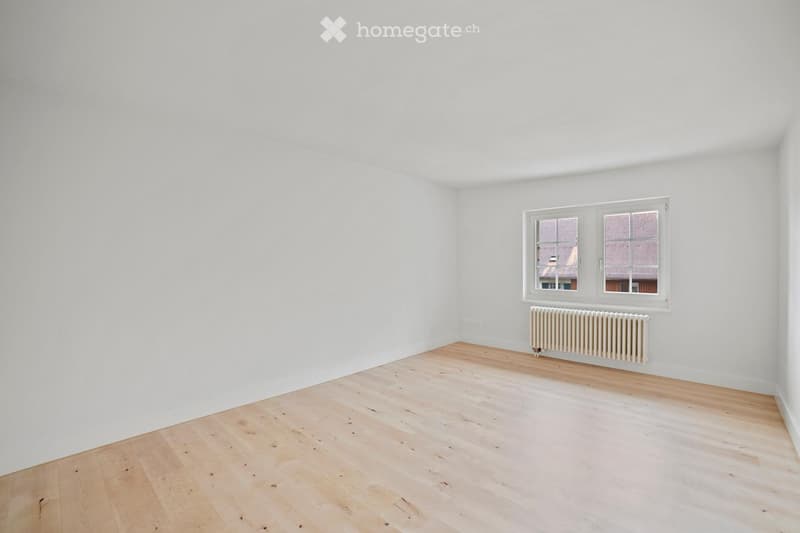 Empty room with white walls, light wooden flooring, and a single window with a radiator below it.