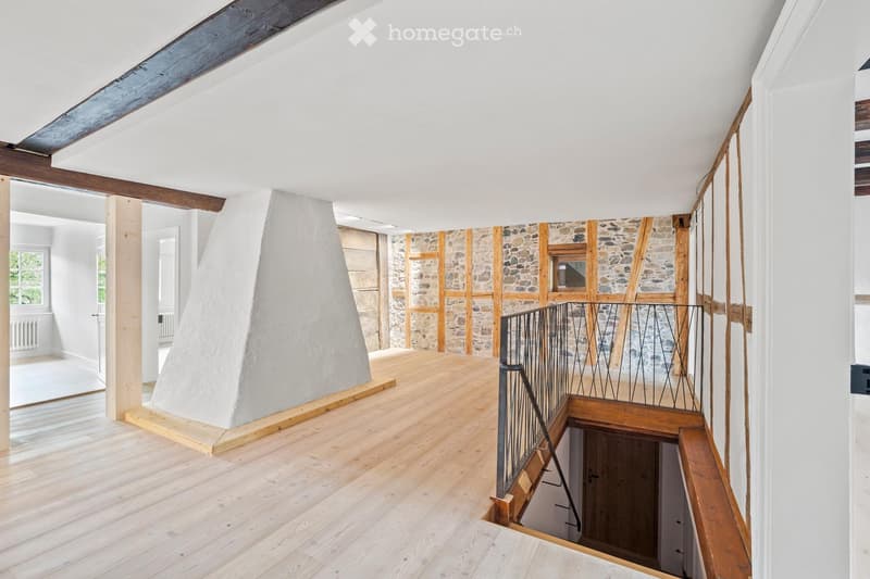 Bright, open room with wooden flooring, exposed wooden beams, and a stone wall section. A modern staircase with a metal railing leads to a lower level.