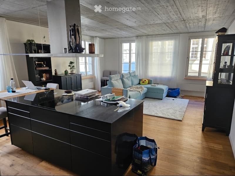 Open-plan living and dining area with a modern kitchen island, light blue sofa, wooden flooring, large windows, and various furniture and decor items.
