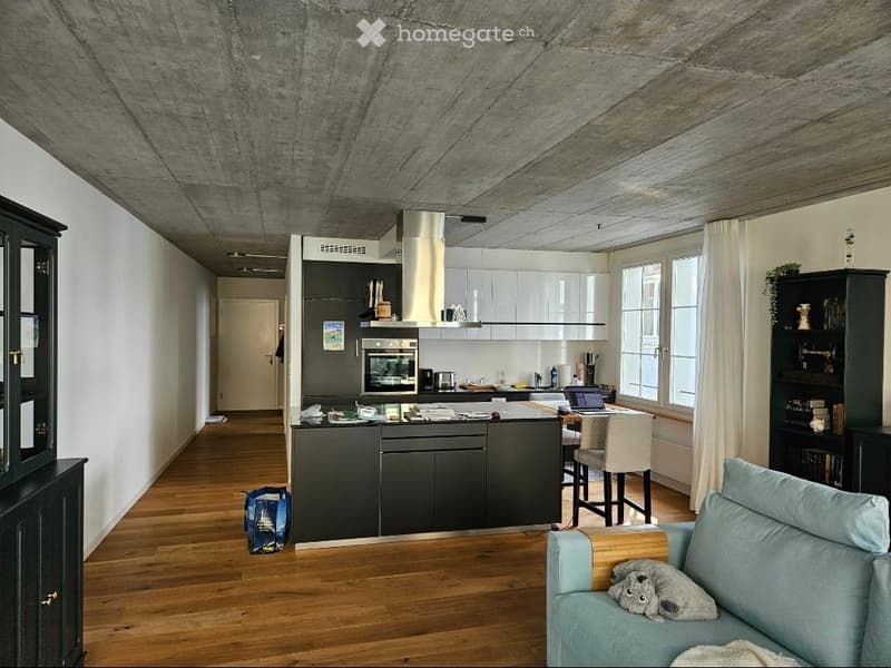Modern open-plan living and kitchen area with wooden flooring, grey sofa, kitchen island, and large windows.