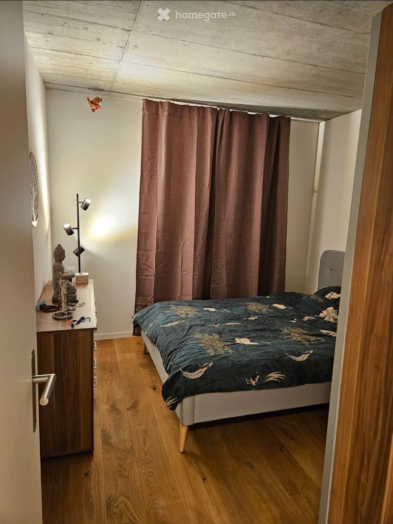 Cozy bedroom with wooden flooring, a bed with a patterned duvet, a nightstand with a lamp, a small table with decorative items, and brown curtains.