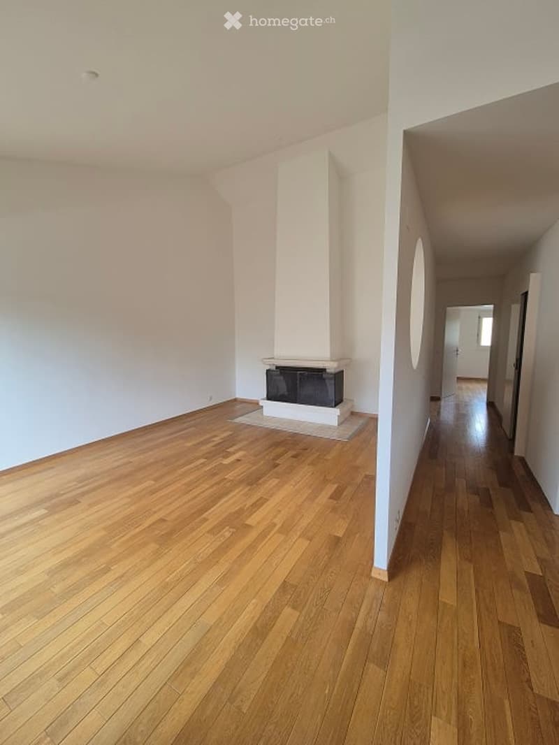 Empty room with wooden flooring, white walls, and a fireplace in the corner. The room is unfurnished and has a hallway leading to another space.