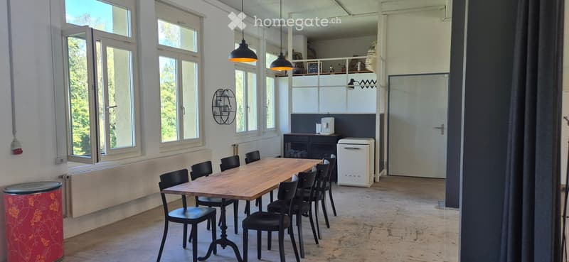 Bright, open dining area with large windows, wooden table, black chairs, small kitchen area with refrigerator and cabinet, and hanging lamps.