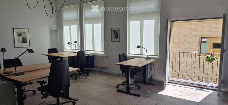 Bright, open-plan office space with large windows, wooden tables, black office chairs, adjustable lamps, and framed artwork on the walls. A balcony with a metal railing is visible through an open doorway.