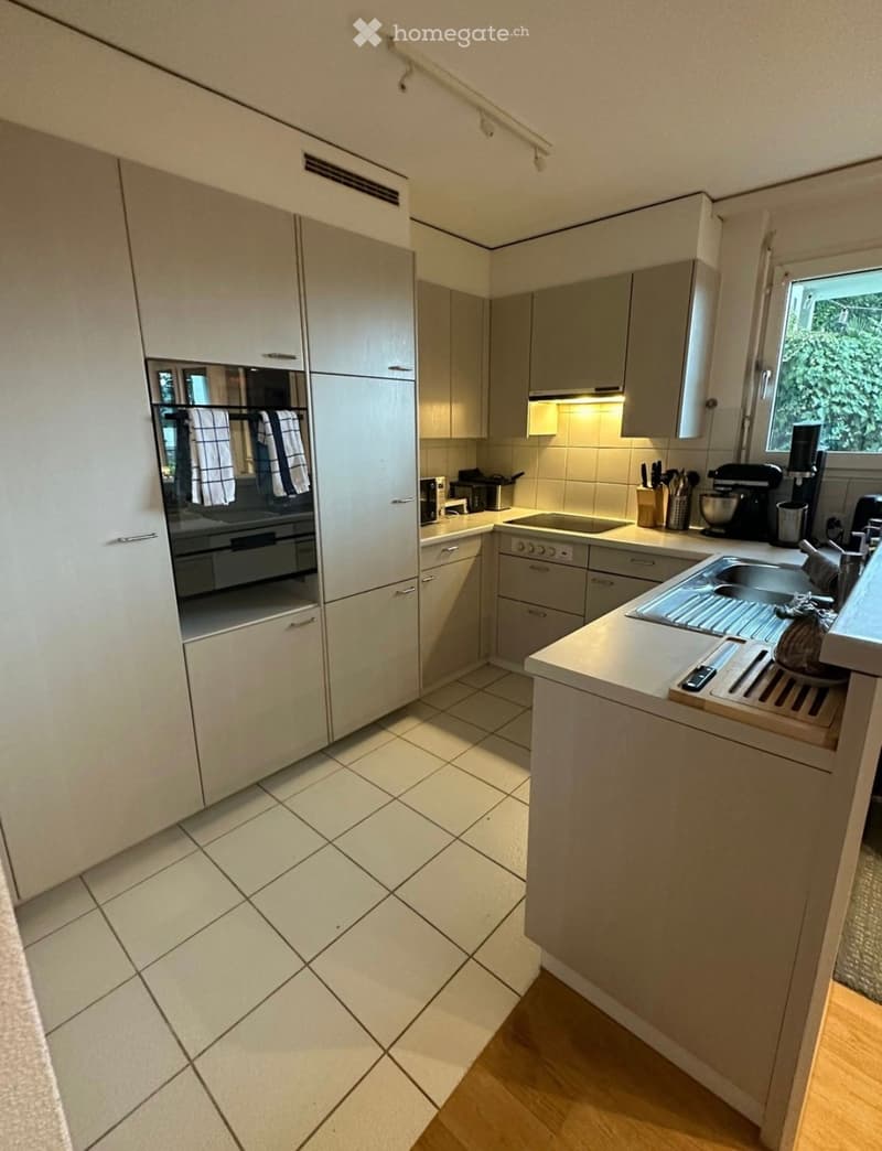 Modern kitchen with white cabinets, tiled floor, and wooden countertop. Features a sink, oven, microwave, coffee maker, and blender.