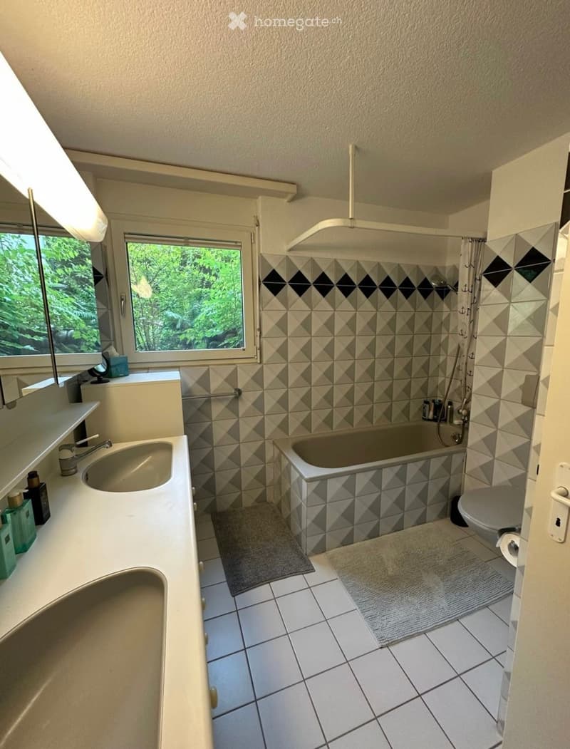 Bathroom with a sink, bathtub, toilet, and shower area. The walls are tiled with a geometric pattern, and there are two rugs on the floor. A window offers a view of greenery outside.