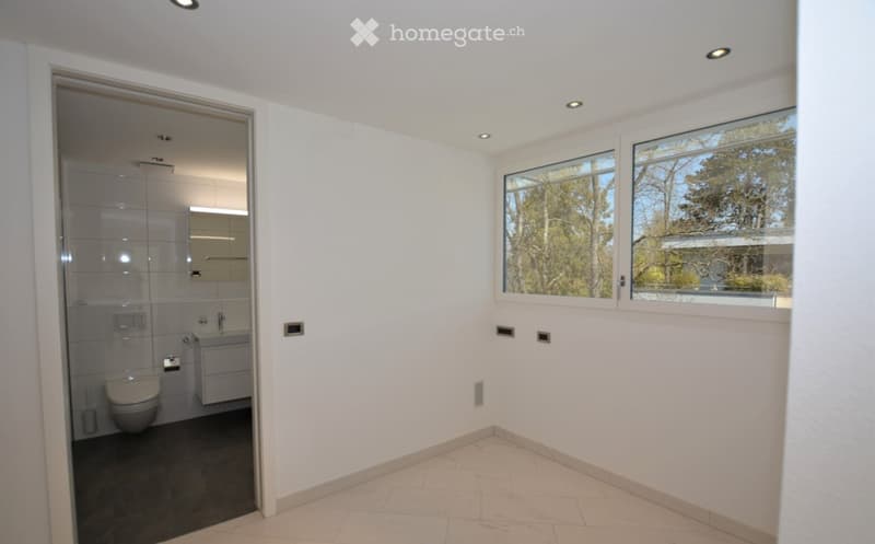 Modern bathroom with white tiled walls, a toilet, a sink, and a glass-enclosed shower. Large windows provide natural light and a view of greenery outside.