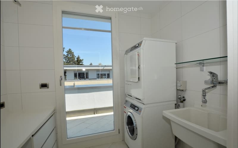 Modern bathroom with white tiles, a sink with a chrome faucet, a washing machine, and a large window offering a view of the outdoor area.