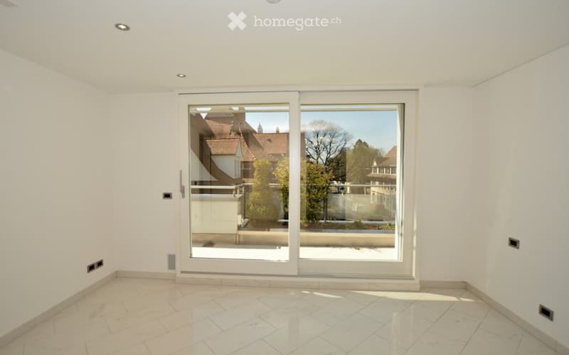 Empty room with white walls, marble tiled floor, and large sliding glass doors leading to a balcony with a view of neighboring houses and trees.