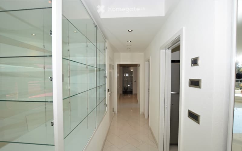 Modern hallway with white walls, recessed lighting, glass partitions, and tiled flooring. Features a small bathroom visible at the end of the corridor.
