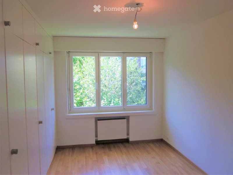Empty room with wooden flooring, white walls, large window with white frame, radiator, and a single ceiling light fixture.