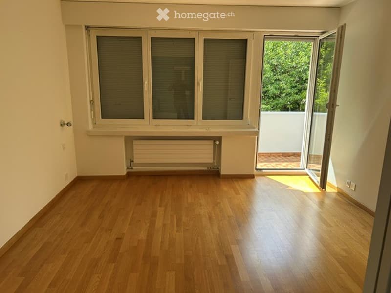 Empty room with wooden flooring, large windows, radiator, and an open door leading to a balcony.