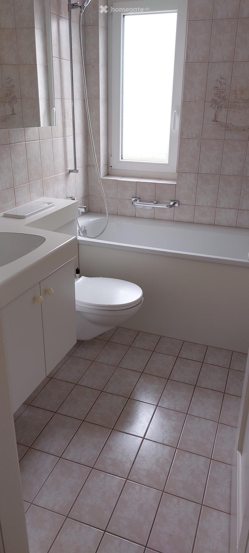 Modern bathroom with beige tiles, a toilet, bathtub, sink, and a window above the bathtub.