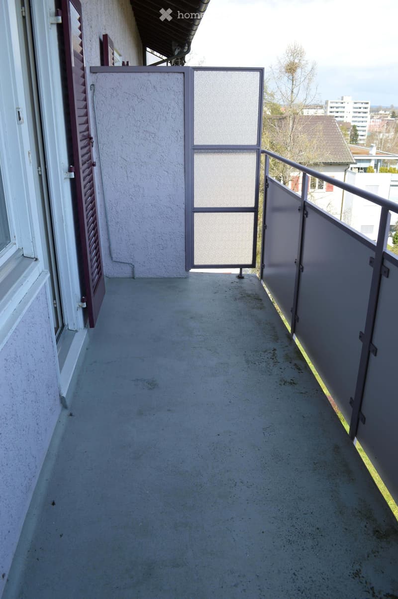 Balcony with concrete floor, metal railing, and a gate leading to the exterior. The balcony is adjacent to a building with white walls and red shutters.