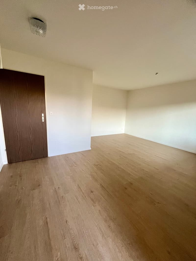 An empty room with wooden flooring and white walls, featuring a door on the left side