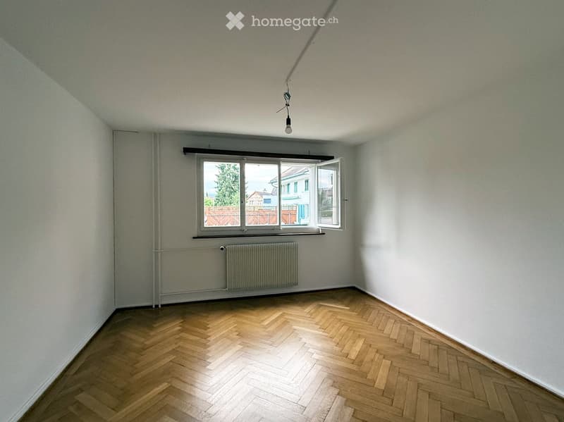 Empty room with wooden herringbone flooring, white walls, a window with open shutters, radiator, and a single ceiling light fixture.