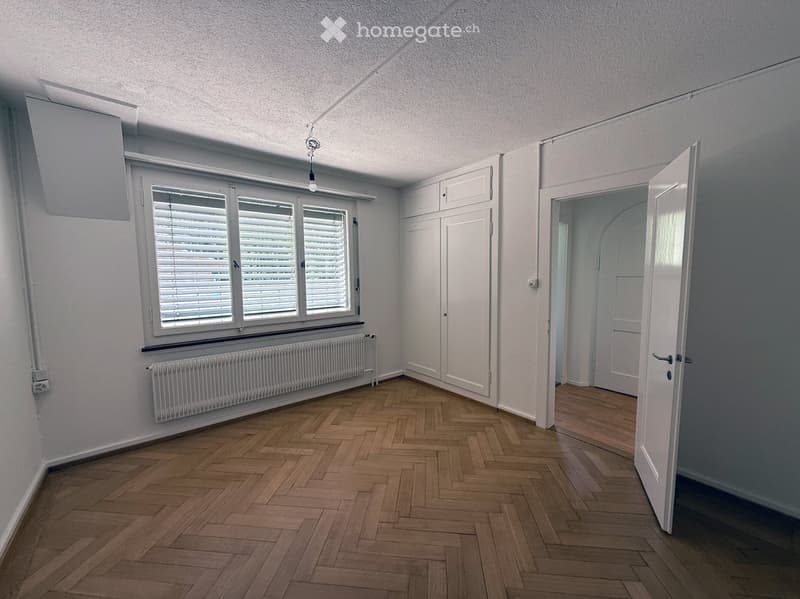 Empty room with wooden herringbone flooring, white walls, large window with blinds, radiator, and open door leading to another space.
